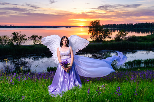 young beautiful white girl with angel wings and flying veil stands on a hillside with violet wildflowers bouquet in her hands.