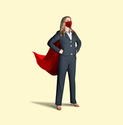A woman stands confidently with her hands on her hips as she wears both a red cape and a red protective face mask isolated on a yellow background.