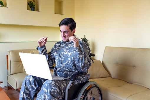 A young soldier in military uniform is sitting on the sofa in his living room and feeling happy while communicating with his family, cousins or friends over his laptop.