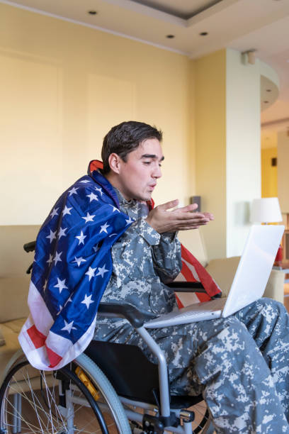 istock Retired Soldier With Physical Disabilities is Using a Laptop and Holding US Flag 1258035733