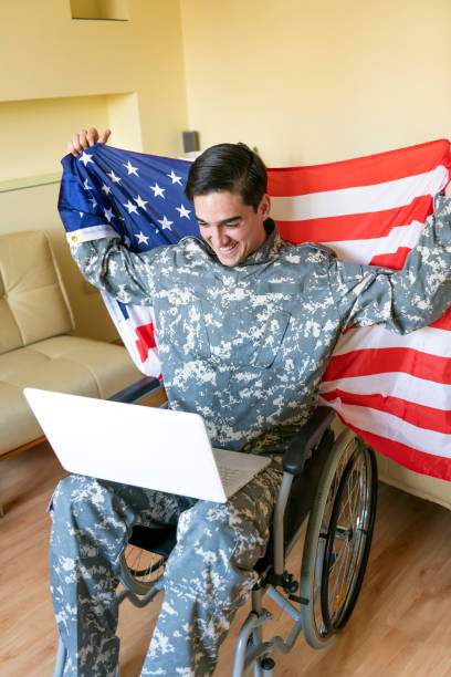 istock Retired Soldier With Physical Disabilities is Using a Laptop and Holding US Flag 1258035785