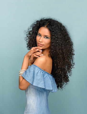 Beautiful fashionable slender young woman from Santo Domingo with long curly black hair wearing subtle makeup posing sideways looking at the camera against a blue studio background