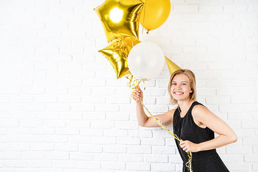 Birthday party. Young blond smiling woman wearing birthday hat holding golden balloons celebrating birthday with copy space