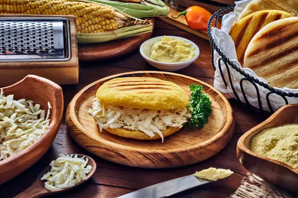 Venezuelan traditional food (Arepa con queso blanco) or yellow corn Arepa with shredded cheese. Ingredients on a table in a rustic kitchen. Corn flour, fresh cheese, corn cobs, butter, water and salt. Arepas, or corn bread, is a main meal in the Venezuelan Culture and cuisine.