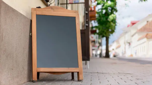 black empty chalkboard in wooden frame stands on pavement near street cafe entrance with open door cityscape background outdoor