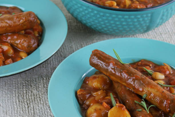 Stock photo showing close-up view of turquoise plates and serving dish of sausage, potato, carrots and bean casserole which has been slowly cooked in a flavoursome tomato sauce. imagen de primer plano de la cazuela de salchicha casera con patatas, zanahorias y frijoles en sabroso salsa de tomate / estofado en platos de color azul turquesa, tazón de azul turquesa sobre fondo de textura gris, se centran en primer plano