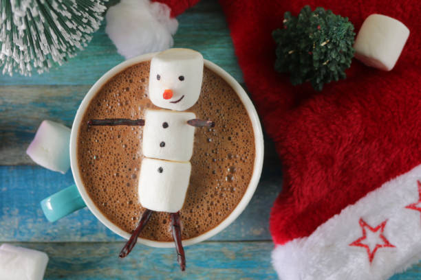 istock Close-up image group of objects, model Christmas trees, mug hot chocolate drink / cocoa with marshmallow snowman, Father Christmas figurine, string fairy light decoration, red and white Santa hat and marshmallows, turquoise tongue groove effect backdrop 1258117077