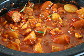 Image of homemade sausage casserole with potatoes, carrots and beans being cooked in slow cooker with fresh herbs, flavoursome tomato gravy / stew elevated view