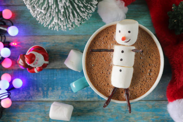 istock Close-up image group of objects, model Christmas trees, mug hot chocolate drink / cocoa with marshmallow snowman, Father Christmas figurine, string fairy light decoration, red and white Santa hat and marshmallows, turquoise tongue groove effect backdrop 1258117133