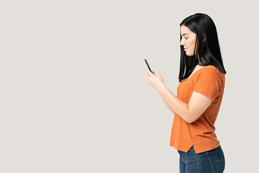 Side view of young woman text messaging through smartphone while standing in studio with gray background