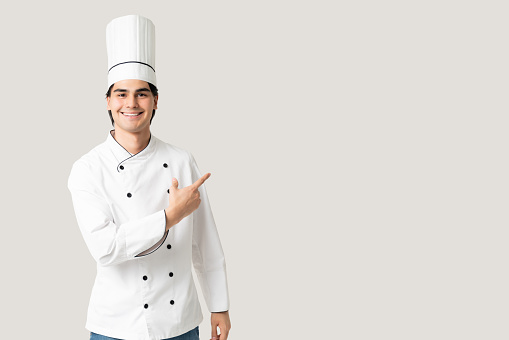 Good looking Hispanic male chef pointing finger towards blank space in studio with gray background