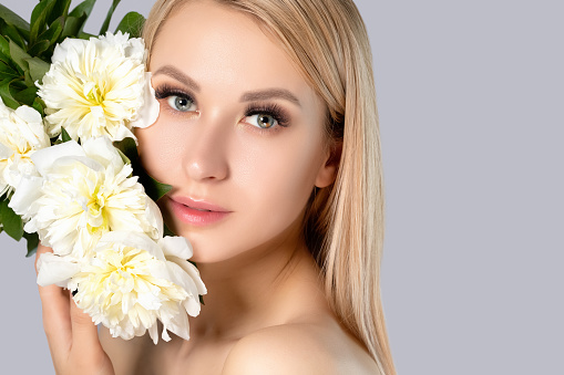 Portrait of a beautiful blonde woman with long hair, beautiful fresh makeup and healthy clean skin with peonies in her hands. Makeup and cosmetology concept.
