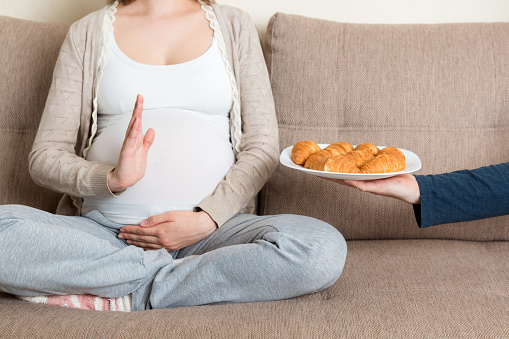 Close up of pregnant woman relaxing on the sofa makes stop gesture to croissants. Expecting mother refuses to eat pastry. Diet during pregnancy concept.