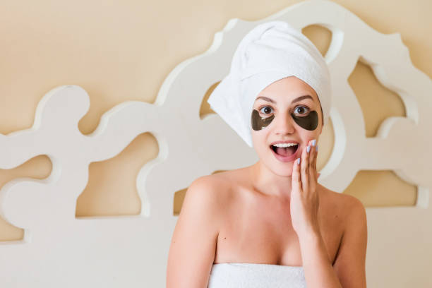 mujer sorprendida envuelta en toalla después de la ducha de la mañana y con manchas negras debajo de los ojos es relajarse en la cama y usar su teléfono. retire las ojeras. concepto de cuidado de la piel - human skin sparse washing face human face fotografías e imágenes de stock