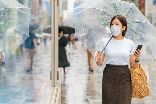 jeune femme retenant le parapluie et le téléphone intelligent tout en marchant dans la ville pendant la pluie - saison des pluies photos et images de collection