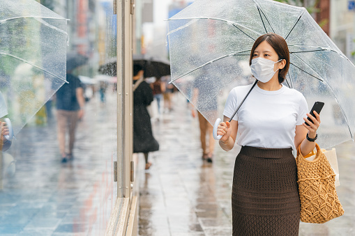 A young woman is holding an umbrella and a smart phone while walking in the city during rain.