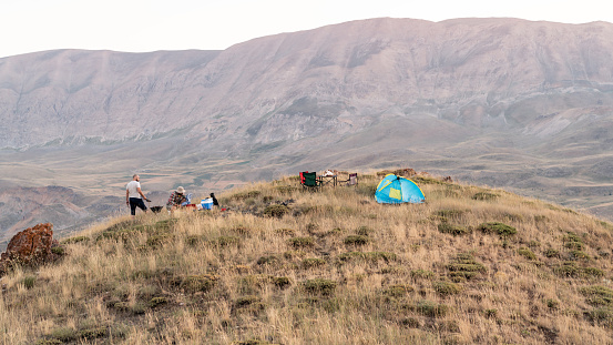 Two friends are camping on top of hill in countryside during summertime. Blue tent is seen on the right side of horizontal frame. Shot under daylight with a full frame mirrorless camera.