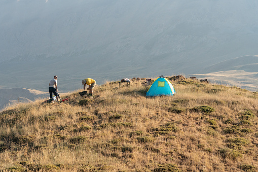 Two friends are camping on top of hill in countryside during summertime. Blue tent is seen on the right side of horizontal frame. Shot under daylight with a full frame mirrorless camera.