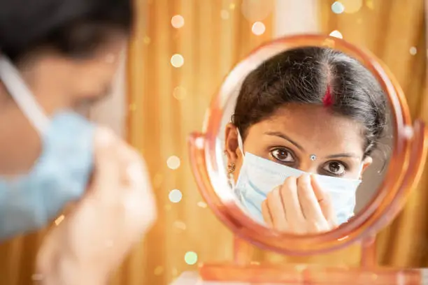 Indian woman getting ready infront of mirror by adjusting medical mask before going out during festival celebration - concept of new normal due to coronavirus or covid-19 pandemic.