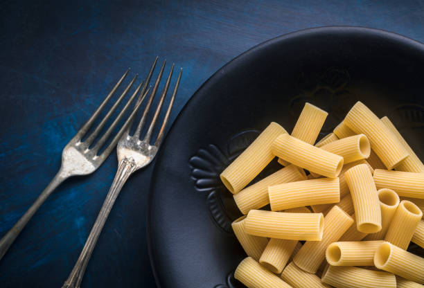 Pasta rigatoni overhead arrangement with two old forks in black plate on blue background studio shot