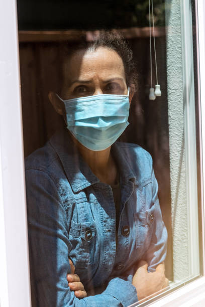 istock Sad mature woman looking through a window wearing a protective face mask 1258166895