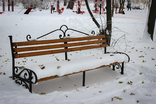 In The Winter Park, An Empty And Lonely Bench Is Completely Covered With Snow. The Snow Is Covered With Leaves That Have Fallen From The Trees.