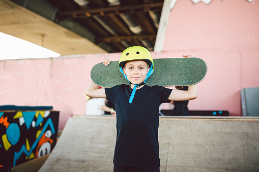 Caucasian child skateboarder posing with skate board on shoulders an outdoor skate park. Portrait of boy is engaged in skateboarding on half pipe ramp. Outdoor activities and extreme sports in summer.