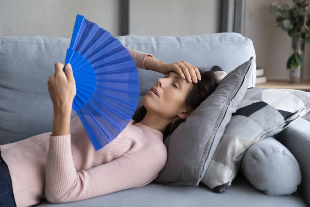 istock Overheated young woman relax on sofa wave with hand fan 1258280095