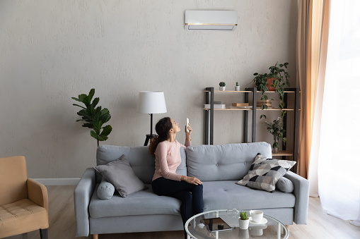 Young Caucasian woman sit on sofa in living room turn air condition device with remote controller, smiling millennial female relax on couch at home switch conditioner, breathe fresh air in apartment