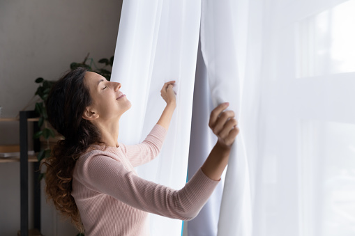 Happy young Caucasian woman open curtains breathe fresh air enjoy early morning sunshine, smiling relaxed millennial female welcome new sunny day, feel optimistic positive, stress free concept