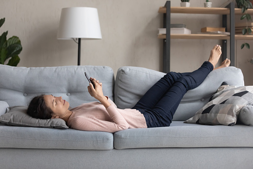 Young caucasian woman lying relaxing on couch in living room browsing wireless internet on smartphone, millennial girl rest on sofa texting messaging on cellphone gadget, enjoy lazy weekend at home