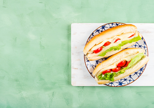 Submarine sandwich made of freshly baked mini baguette with tomatoes, lettuce and melted cheese on white wooden board over green background. Quick healthy lunch