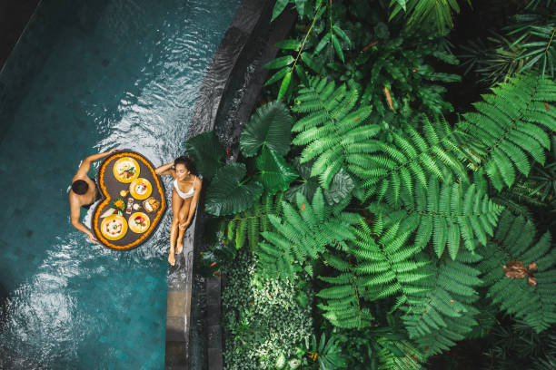 Travel happy couple in love eating floating breakfast in jungle swimming pool. Awakening in morning. Black rattan tray in heart shape, Valentines day or honeymoon surprise, view from above. stock photo