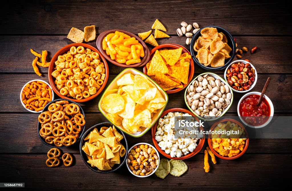 Salty snacks assortment shot from above on rustic wooden table Party food: assortment of salty snacks in bowls shot from above on wooden table. The composition includes potato chips, popcorn, corn bugles, pretzels, peanut, pistachio, cheese sticks, nachos and salsa and others. Top view. Predominant colors are yellow and brown. High resolution 42Mp studio digital capture taken with SONY A7rII and Zeiss Batis 40mm F2.0 CF lens Potato Chip Stock Photo