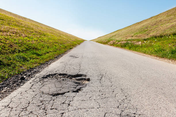 istock Abandoned asphalt road with big pothole in foreground leading into distance, surrounded with green hills on both sides 1258687701