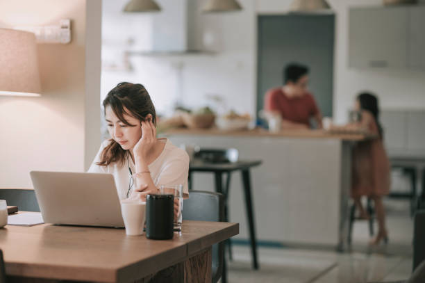 una asiática china media mujer adulta que trabaja en el comedor escribiendo usando su computadora portátil mientras su marido preparaba la comida en la cocina con su hija - malaysian person family asian ethnicity mother fotografías e imágenes de stock