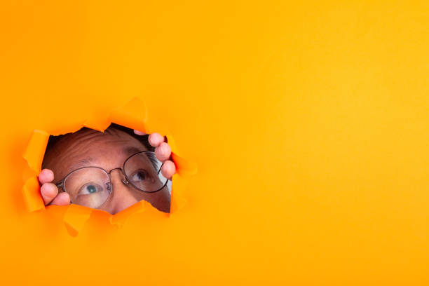istock The man with spectacle peeks through a cut hole in the orange paper background. 1258733806