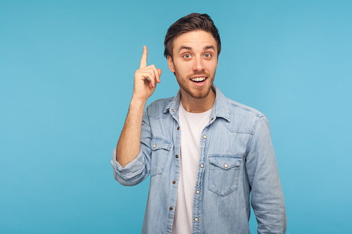 Portrait of happy inspired man in worker denim shirt raising finger and having genius idea, looking amazed by sudden smart solution, creative thought. indoor studio shot isolated on blue background