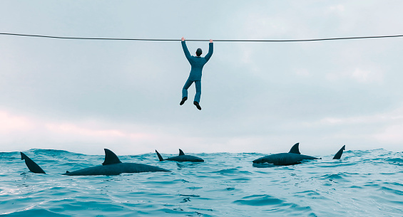 Man holding on to wire across an ocean. Sharks are in the water below him Concept of a situation with great risk where a person needs to find a solution and get to safety fast.  
Note: The man is a 3D-render. Property release attached.