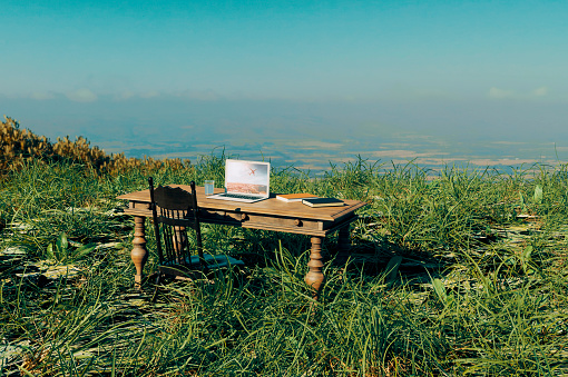 Concept of working from anywhere if you just have your laptop with you. Table and chair stands in the middle of a field. There is a computer on the table and some books and glass of water. 
Note: The image on the computer screen is my work. The image is already on istock: 90702703