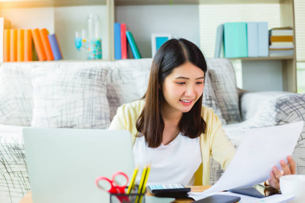 istock Young asian woman working from home on laptop computer and looking paperwork. Beautiful businesswoman working on laptop computer while sitting at living room with smile face. Work from home concept. 1258908169