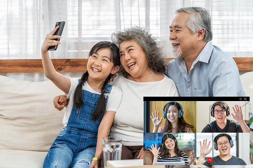 Online meeting, Social distancing concept, Asian Granddaughter with couple Grandparent taking video call conference to Business people saying hello with teamwork colleague by mobile phone in house