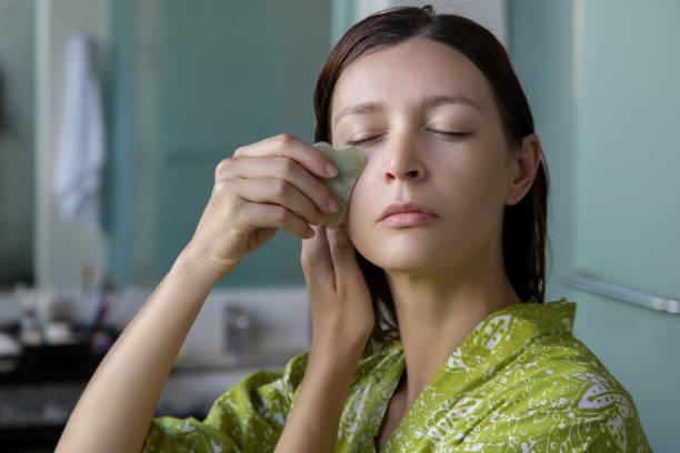 a jovem está fazendo massagem facial gua sha no banheiro em frente ao espelho. pedra de jade gua sha em tratamento domiciliar. - lastone therapy massaging spa treatment stone - fotografias e filmes do acervo