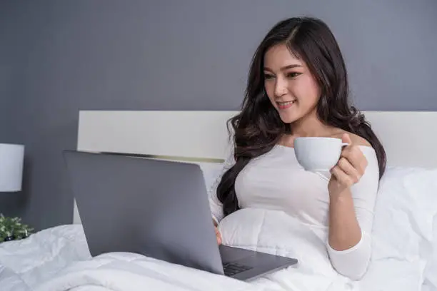 woman drinking a cup of coffee and using a laptop computer on bed