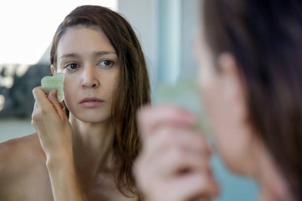 a jovem está fazendo massagem facial gua sha no banheiro em frente ao espelho. pedra de jade gua sha em tratamento domiciliar. - lastone therapy massaging stone shiatsu - fotografias e filmes do acervo