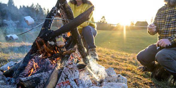 Unrecognizable couple sitting at a campfire and baking marshmallows on a stick. Setting sun in the background. Lens flare.