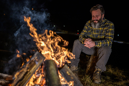 Bearded mid adult man in a checkered shirt sitting on a log next to a campfire and looking at it.