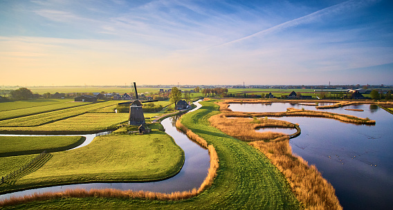 Dutch landscape during sunrise. The picture is made from above. In the middle of the picture is a windmill. There are different canals and between the parts of water are pieces of land consisting of grass.