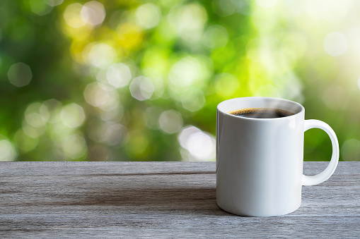 Hot white coffee cup on wooden tabletop on blurred green nature bokeh background