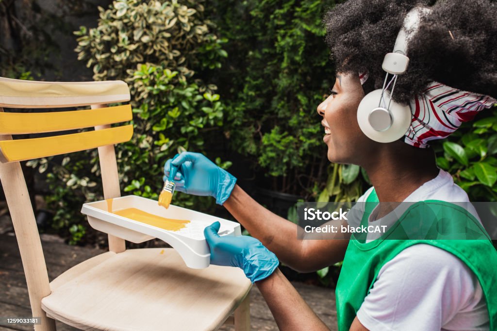 African American woman is painting a chair at home A young woman is painting vintage chair in yellow color with a paintbrush in the back yard while listening to music via wireless headphones DIY Stock Photo